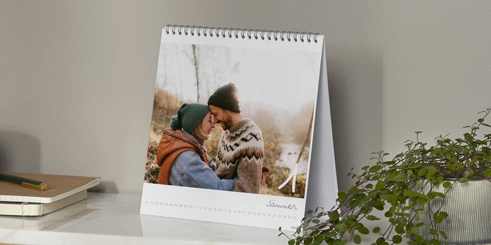 Ein glückliches Paar steht nahe beieinander und umarmt sich, während sie sich liebevoll anlächeln. Auf dem Tisch steht ein CEWE Fotokalender.