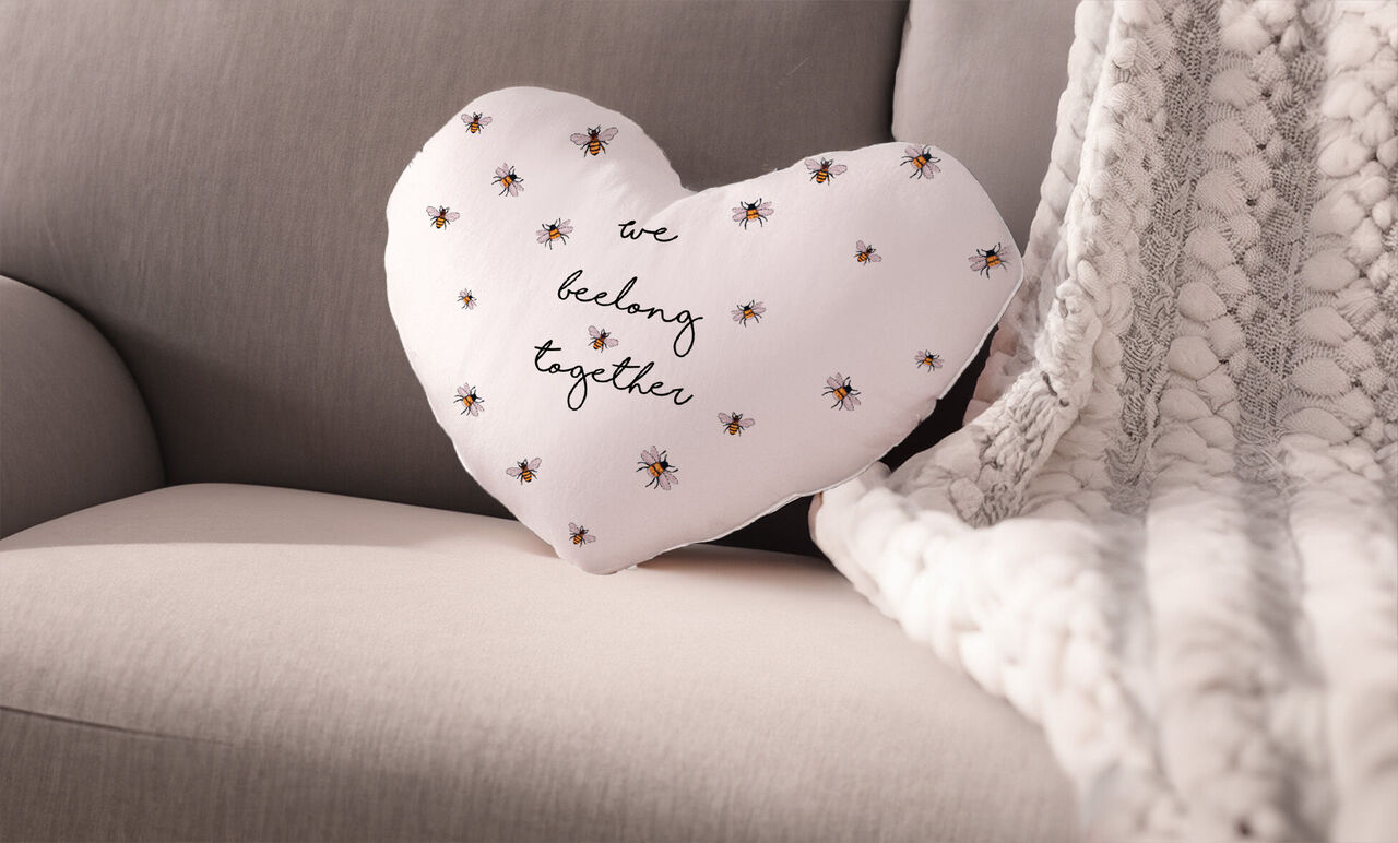 Weißes, herzförmiges Kissen mit dem Aufdruck "we belong together" und kleinen Bienenmotiven, auf einem grauen Sofa.