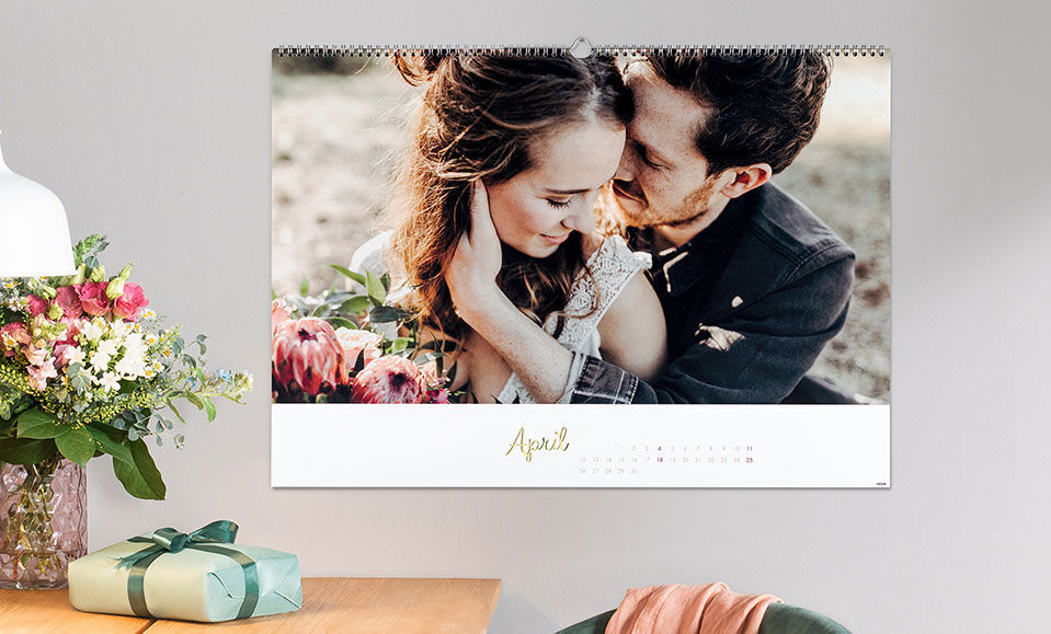 Ein liebevoller Kalender hängt an der Wand mit einem Paar, das sich umarmt und Blumen im Hintergrund. Der Monat April ist gut zu sehen.