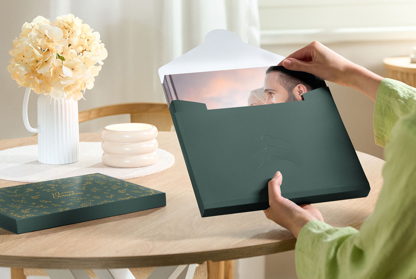 Eine Person öffnet einen grünen Karton mit einem Fotobuch bei einem Tisch. Darin ist ein wunderschönes Bild sichtbar, umgeben vonapril Blumen und kreativem Design.