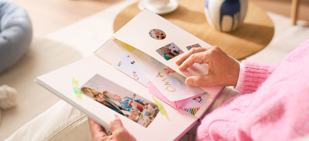 Eine Person blättert in einem CEWE Fotobuch mit bunten Seiten und Fotos von Familienmomenten. Die Person hält das Buch liebevoll in der Hand