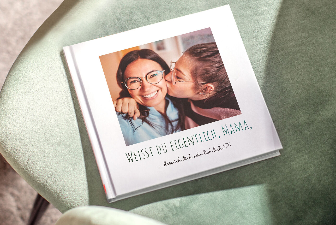Ein liebevolles Fotobuch liegt auf einem gemütlichen Sessel. Auf dem Cover steht „WEISST DU ELGENTILCH, MAMA“.