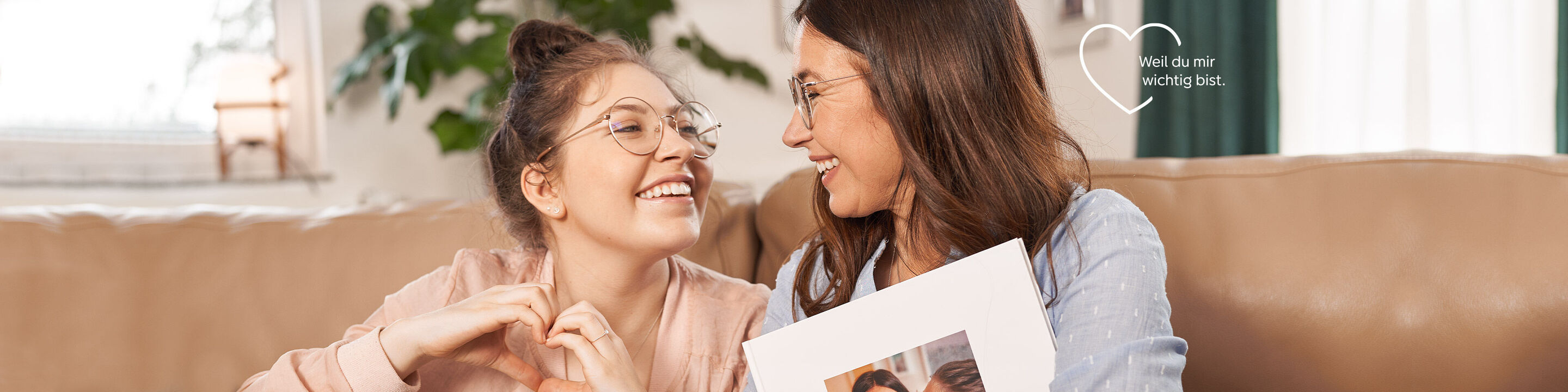 Zwei Frauen sitzen zusammen auf einem Sofa und halten ein Fotobuch in den Händen. Sie teilen einen freudigen Moment und zeigen herzliche Gesten.