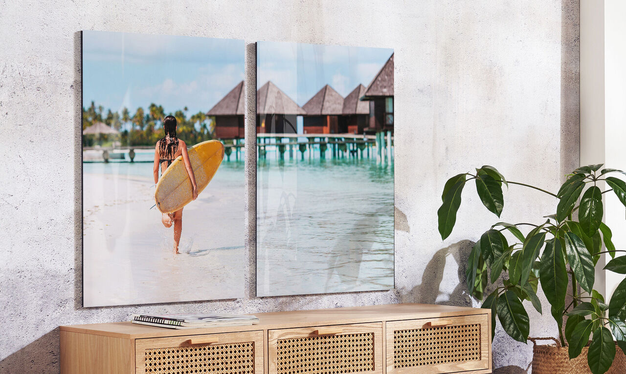 Eine Person trägt ein Surfbrett und geht am Strand spazieren. Im Hintergrund sind Strandvillen und Palmen zu sehen.