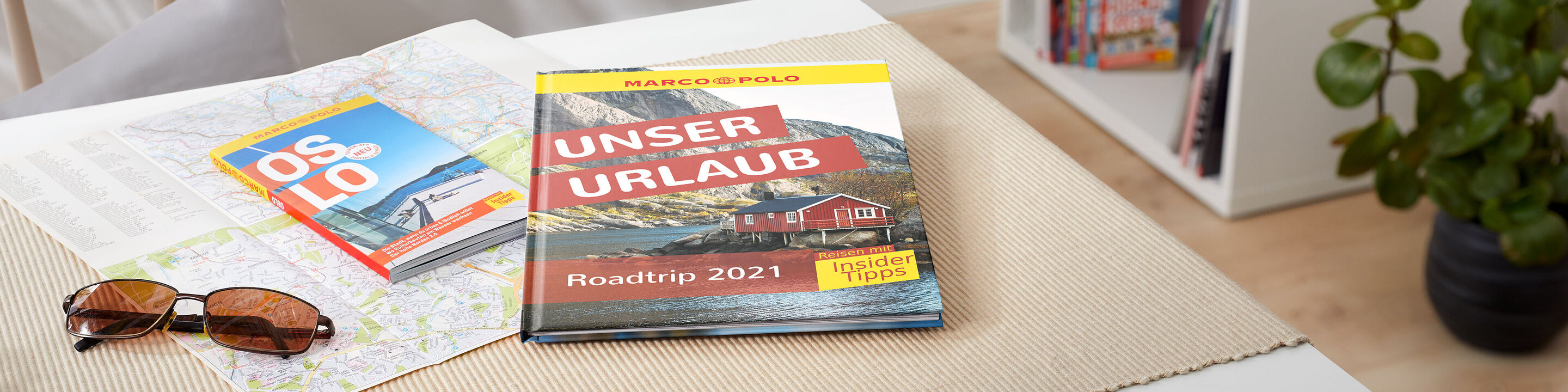 Ein CEWE Fotobuch mit dem Titel 'UNSER URLAUB' liegt auf einem Tisch, umgeben von einer Karte und Sonnenbrille. Der Hintergrund zeigt eine helle, freundliche Raumgestaltung.