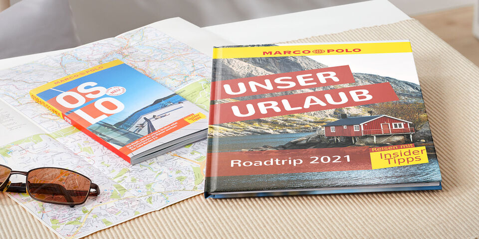 Ein liegendes Fotobuch mit dem Titel 'Unser Urlaub' und einer Karte auf einem Tisch. Dazu liegen Sonnenbrillen und ein Reiseführer.