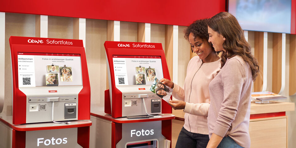 Zwei Personen nutzen einen CEWE Fotoautomaten zum Drucken.