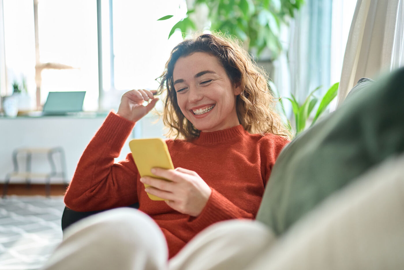 Eine Person entspannt auf einem Sofa und schaut auf ihr Smartphone während sie in einem gemütlichen Raum sitzt. Pflanzen und ein Laptop sind im Hintergrund.