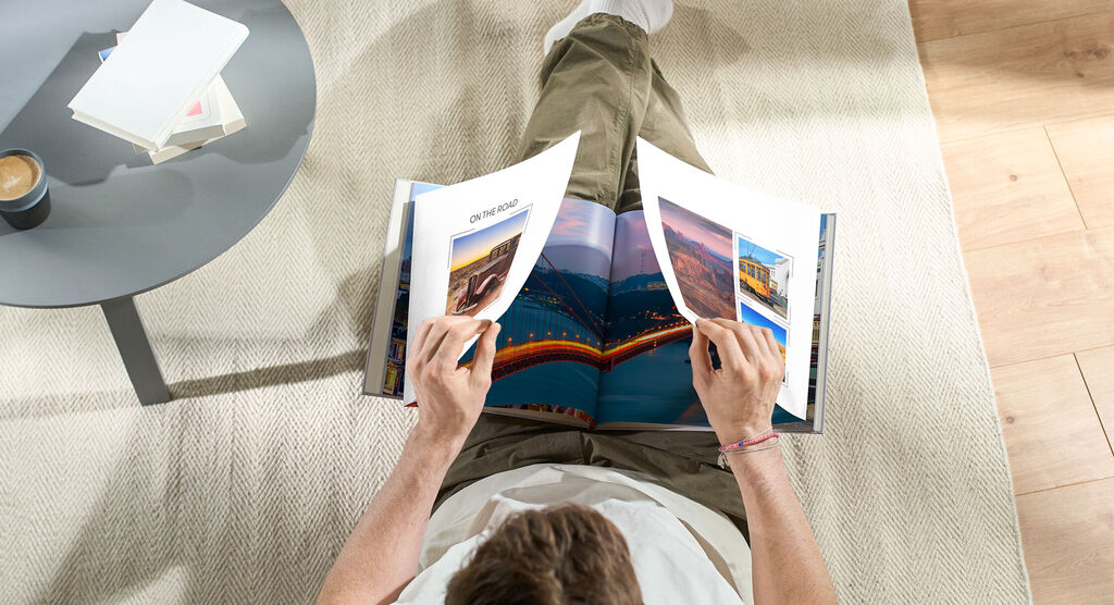 Eine Person blättert in einem offenen Fotobuch auf dem Boden, umgeben von einem Tisch mit einer Tasse.