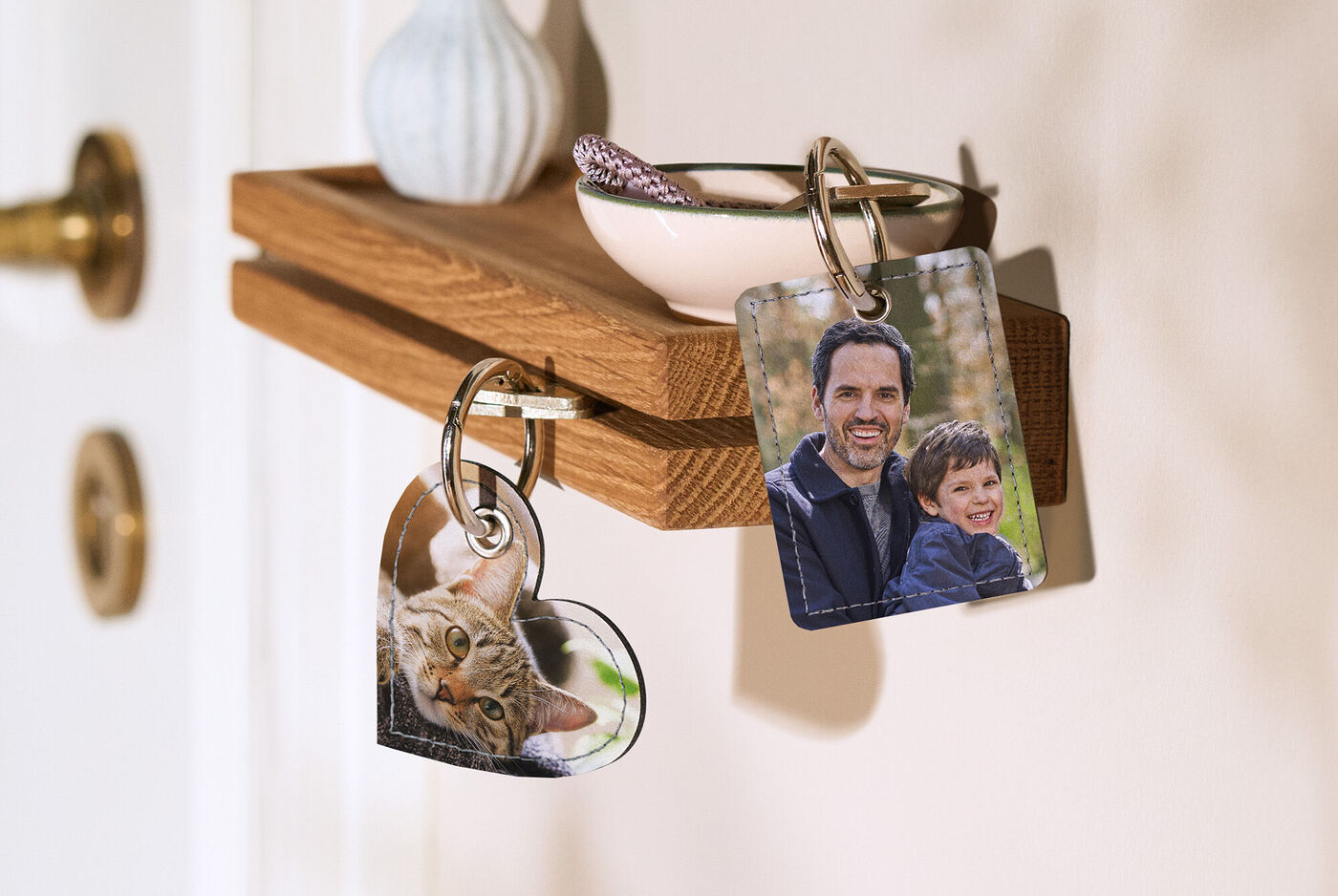 Zwei Schlüsselanhänger hängen an einem Holzregal. Einer hat die Form eines Herzens mit einem Katzenfoto, der andere zeigt zwei Personen in einem schönen Wald. Eine kleine Schüssel und eine dekorative Vase stehen ebenfalls auf dem Regal.