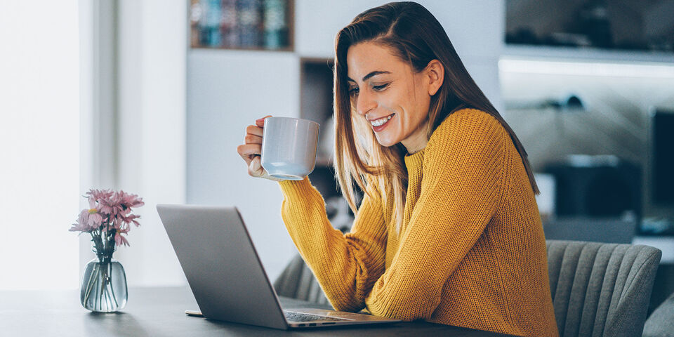 Eine Person mit einem Kaffeebecher sitzt an einem Tisch und arbeitet an einem Laptop. Auf dem Tisch steht eine kleine Blumenvase mit Blumen.
