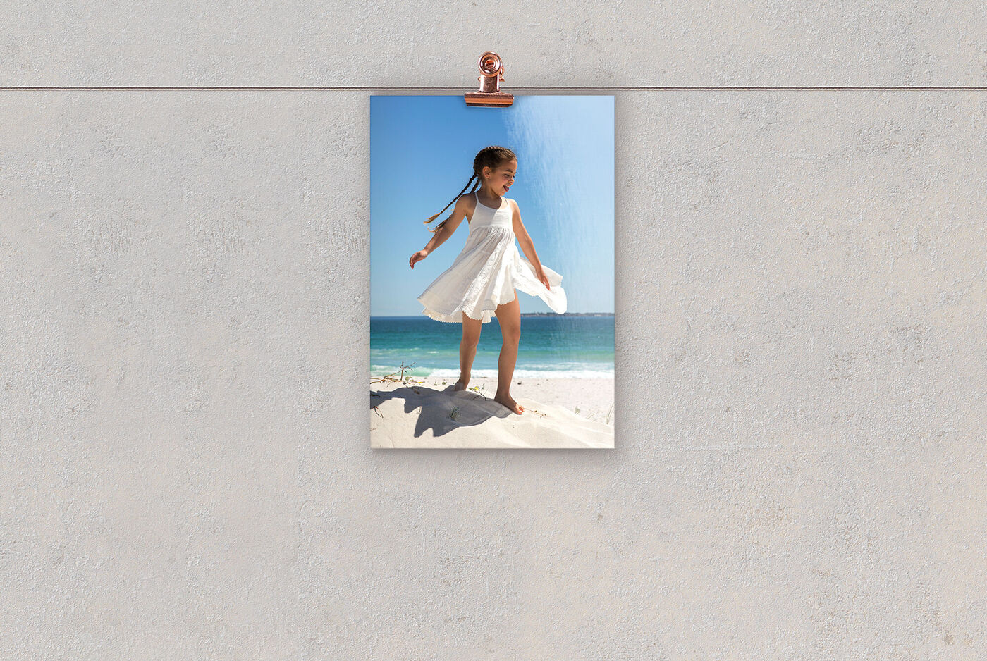 Ein fröhliches Mädchen in einem weißen Kleid steht auf einem Sandhügel am Strand. Hinter ihr ist das blaue Meer und der Himmel zu sehen.