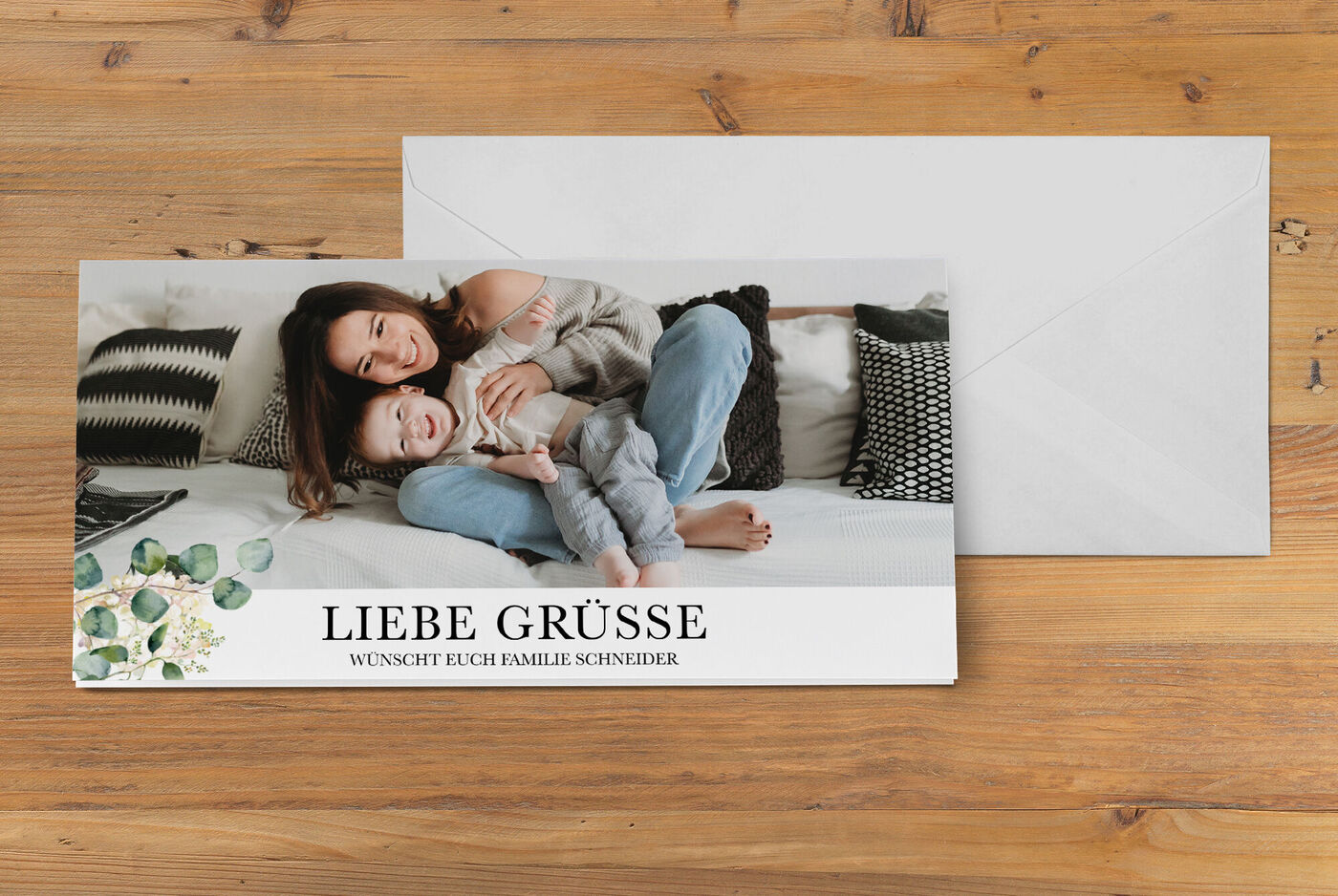 Eine liebevolle Familie posiert auf einem Sofa mit einem fröhlichen Kind, das auf dem Bauch liegt. Die Grußkarte mit dem Text "Liebe Grüße" liegt auf dem Tisch.