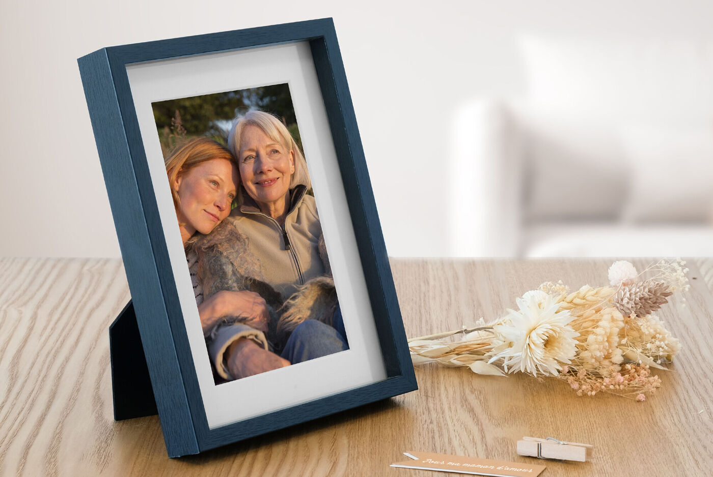 Ein liebevoll gestalteter Rahmen mit einem Foto von zwei lächelnden Personen, auf einem Tisch mit Trockenblumen.