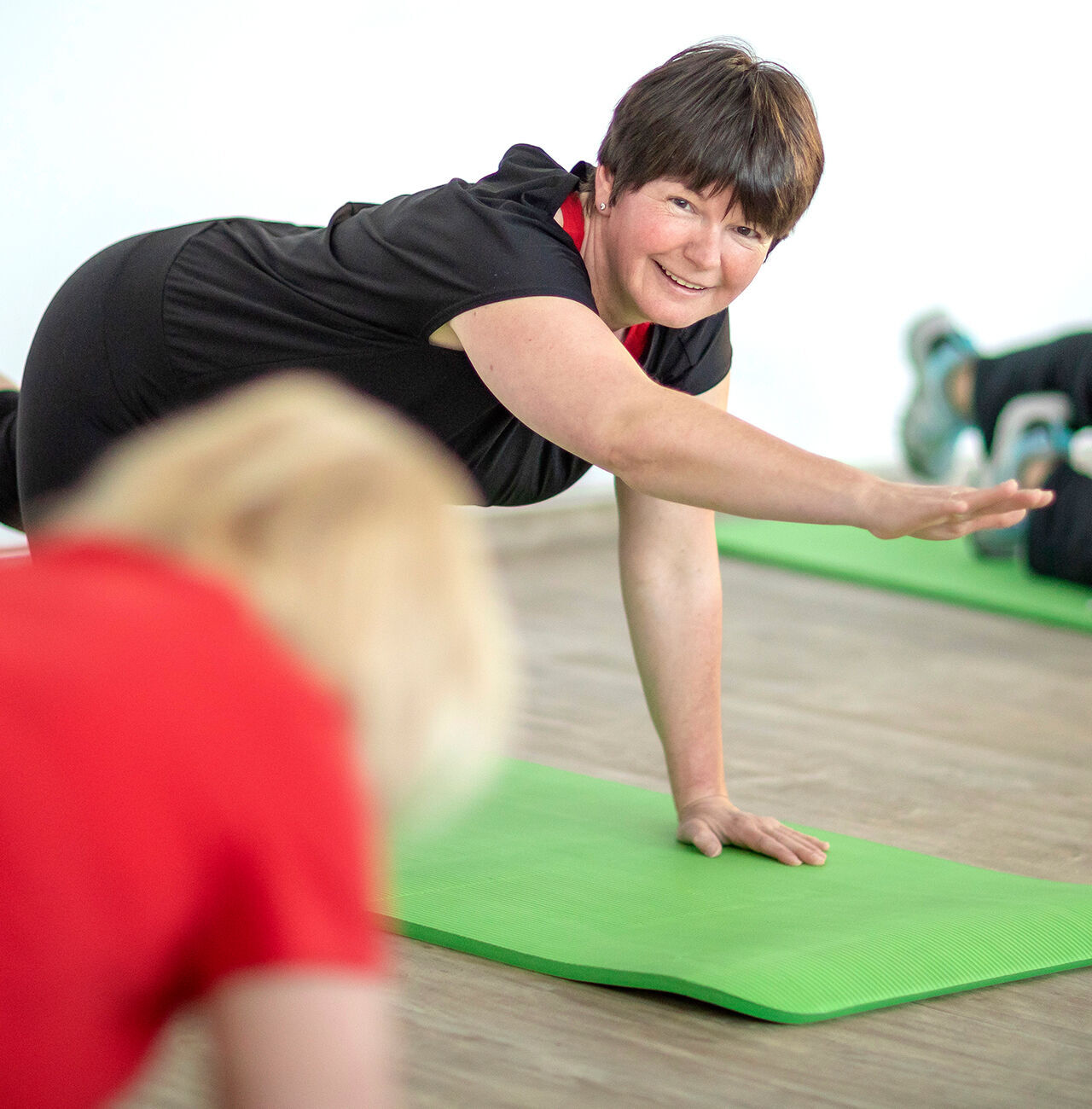 Ein Fitnesskurs mit Teilnehmerinnen auf grünen Matten. Eine Person zeigt eine Übung mit gestrecktem Arm.