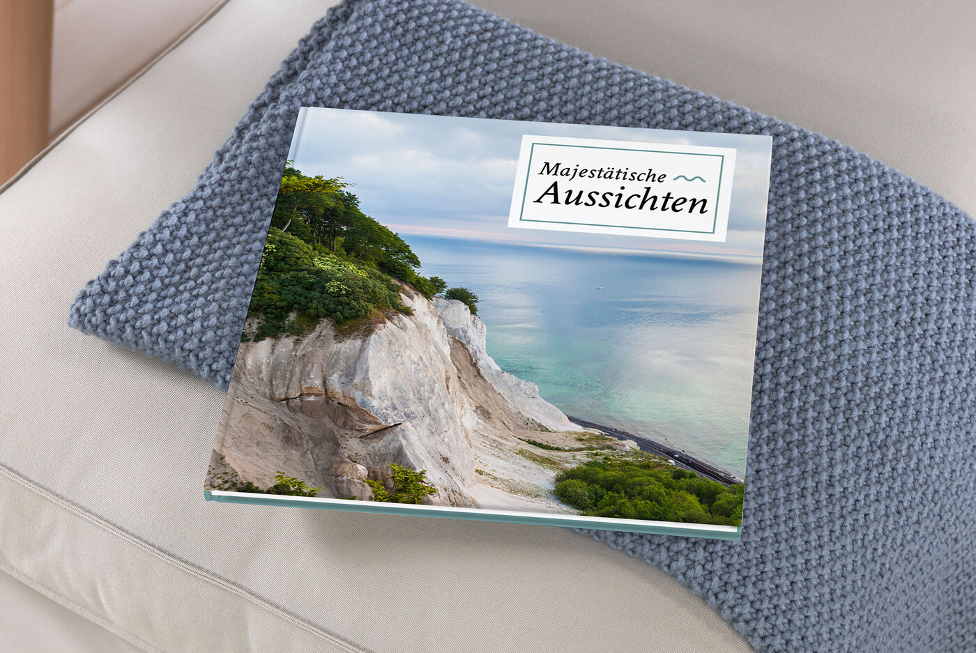 Ein CEWE FOTOBUCH liegt auf einem grauen Kissen. Der Titel lautet "Majestätische Aussichten". Im Hintergrund sieht man eine Küstenlandschaft mit grünem Bewuchs und klarem Wasser.