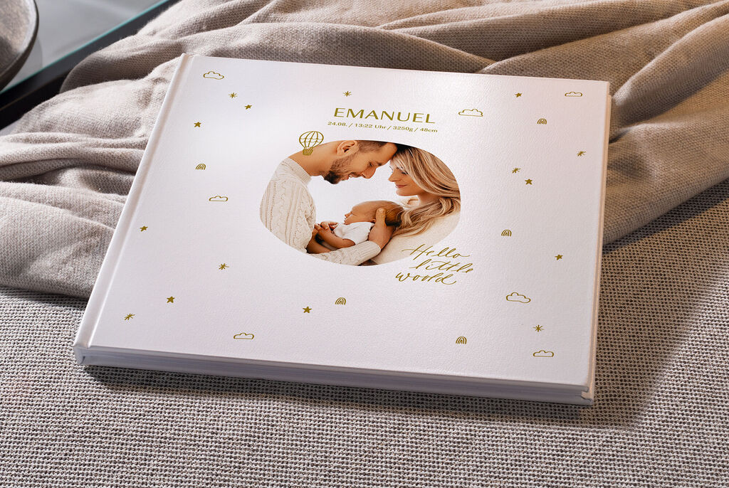 Ein liebevoll gestaltetes CEWE Fotobuch mit dem Namen Emanuel. Das Cover zeigt ein Bild von einem Paar, das glücklich zusammensteht und ein Baby hält. Um das Bild herum sind zarte, goldene Sterne und Wolken.