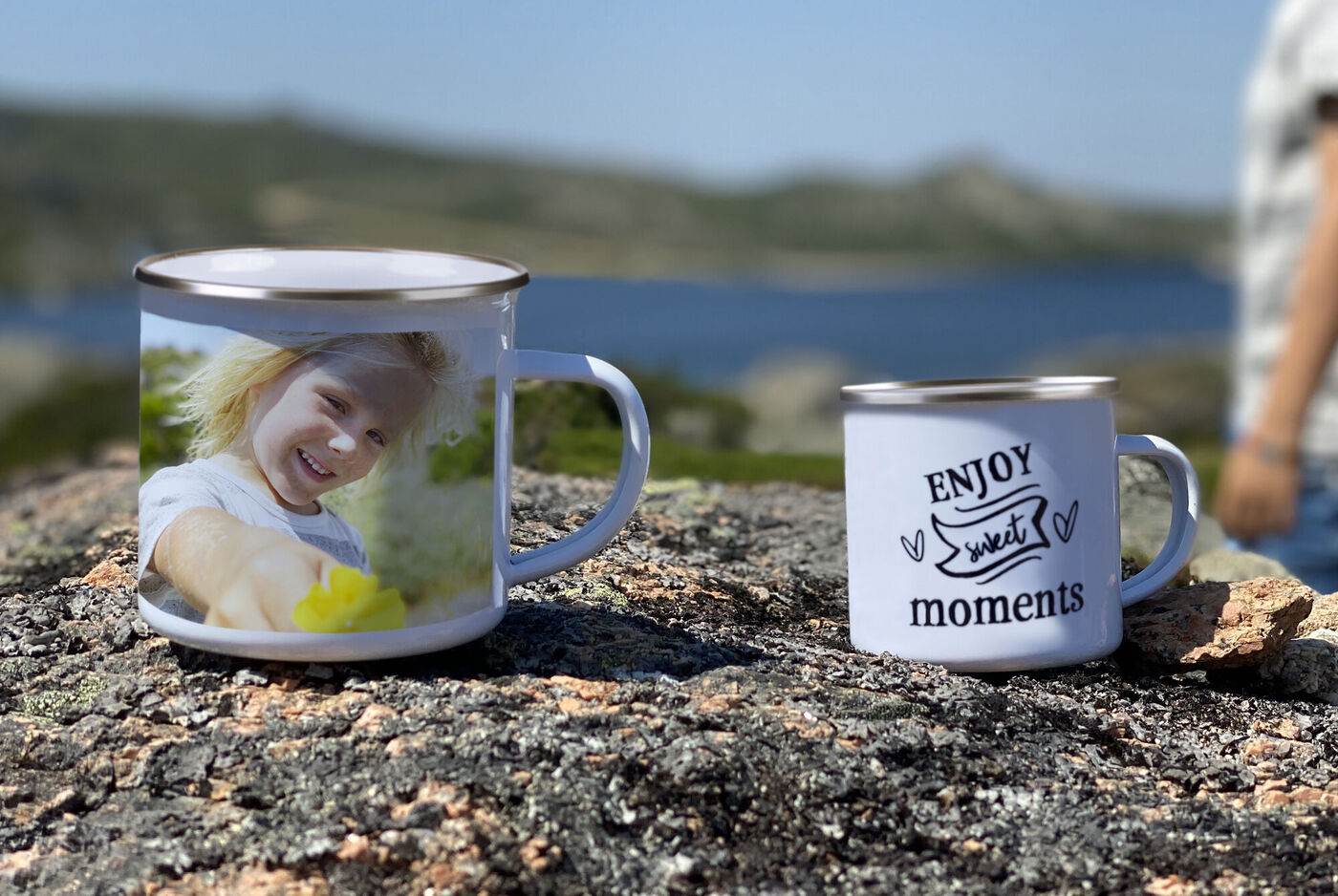 Zwei Tassen auf einem Felsen, mit schöner Aussicht im Hintergrund. Die eine Tasse zeigt ein lächelndes Kind und die andere hat den Schriftzug 'ENJOY sweet moments' darauf. Perfekt für unvergessliche Erlebnisse.