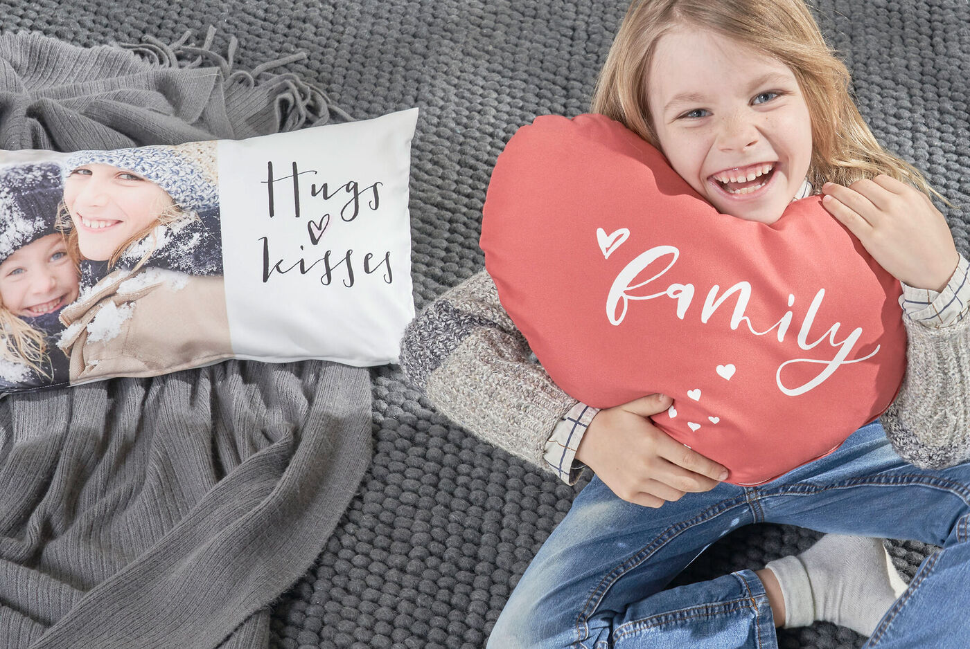 Eine Person sitzt auf einem Teppich und hält ein herzförmiges Kissen mit dem Schriftzug „family”. Links liegt ein Kissen mit dem Text „Hugs & Kisses”.