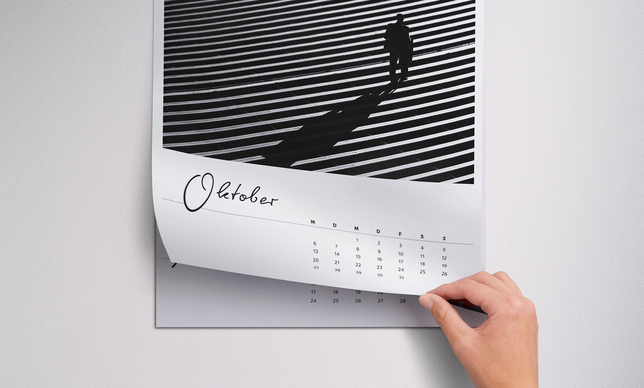 Eine Hand blättert um die Seite eines Fotokalenders mit schwarz-weißen Linien und einer Person im Schatten. Der Monat Oktober ist sichtbar.