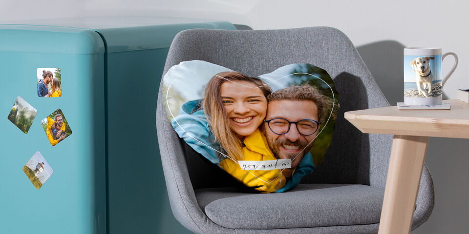 Herzförmiges Kissen mit einem Bild, auf einem Sessel. Eine Tasse und ein Kühlschrank sind im Hintergrund zu erkennen