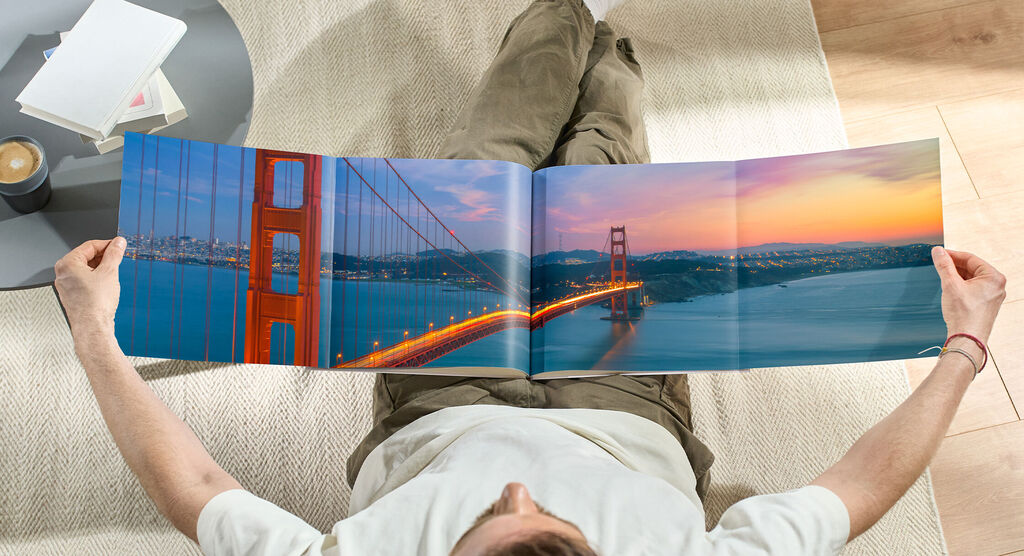Ein Mensch sitzt gemütlich auf dem Boden und betrachtet ein aufgeschlagenes Fotobuch mit einer wunderschönen Fotografie der Golden Gate Bridge bei Sonnenuntergang.