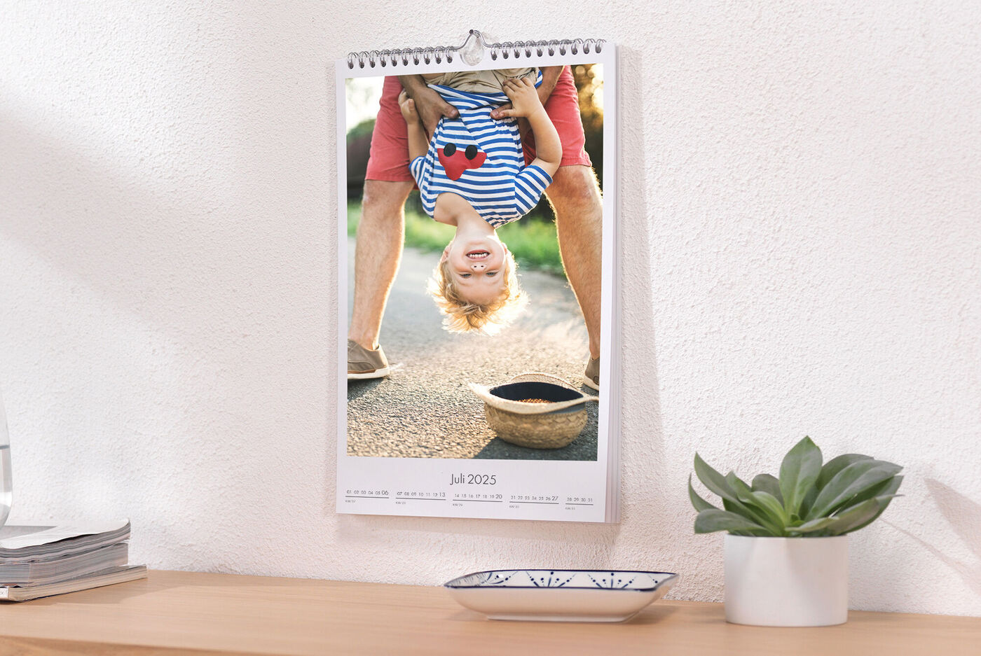 Ein Kind hängt spielerisch kopfüber an einem Tisch. Ein fröhliches Lächeln auf dem Gesicht zeigt Spaß und Lebensfreude. Der Kalender hängt an der Wand über einem Holztisch.