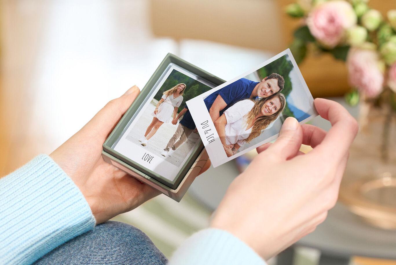 Eine Person hält ein Geschenk mit Fotos von einem Paar in der Hand, umgeben von einem Blumentopf