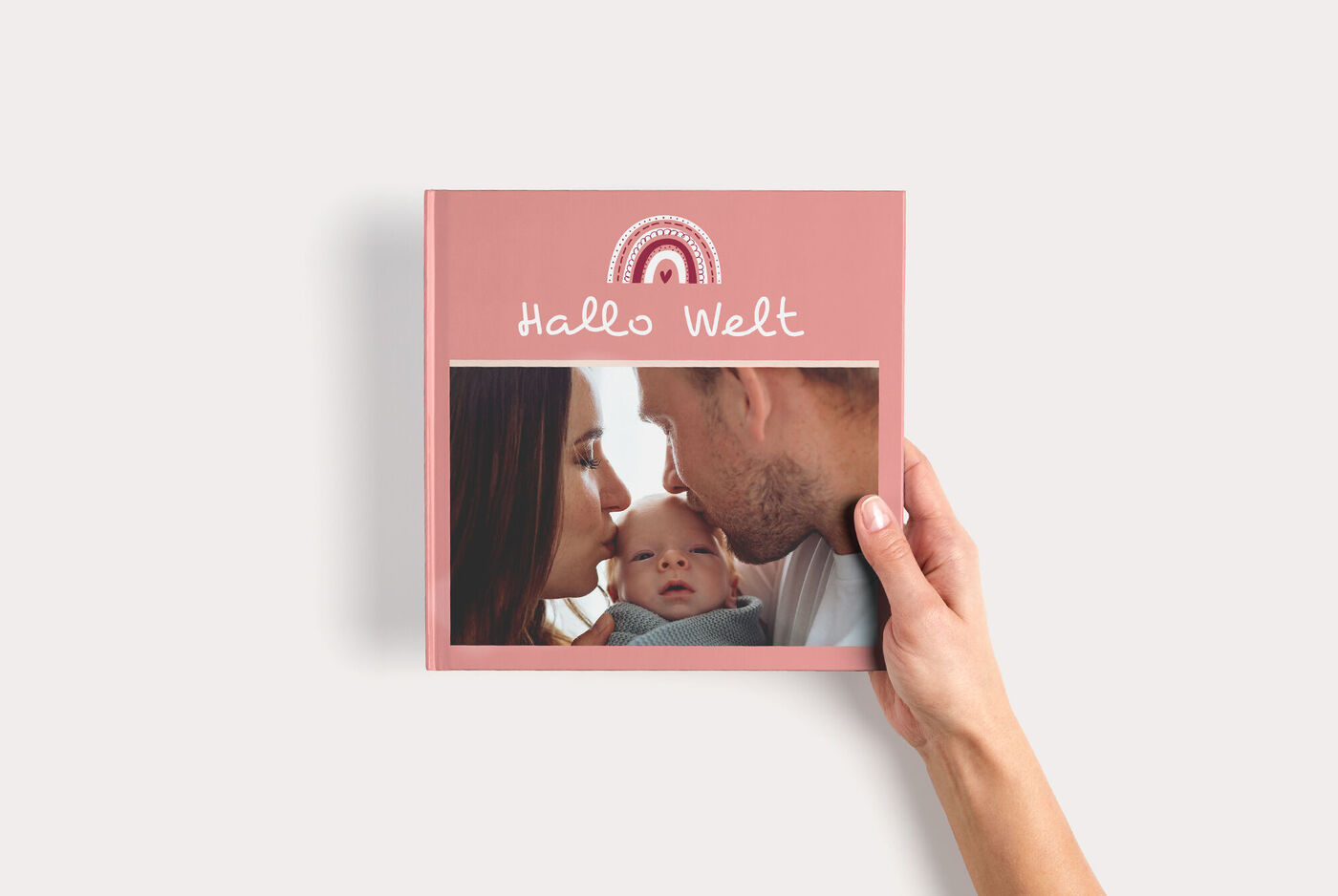 Hand hält ein rosa CEWE FOTOBUCH Quadratisch zum Thema Baby, mit Regenbogenmotiv, Fotofenster und Titel "Hallo Welt"